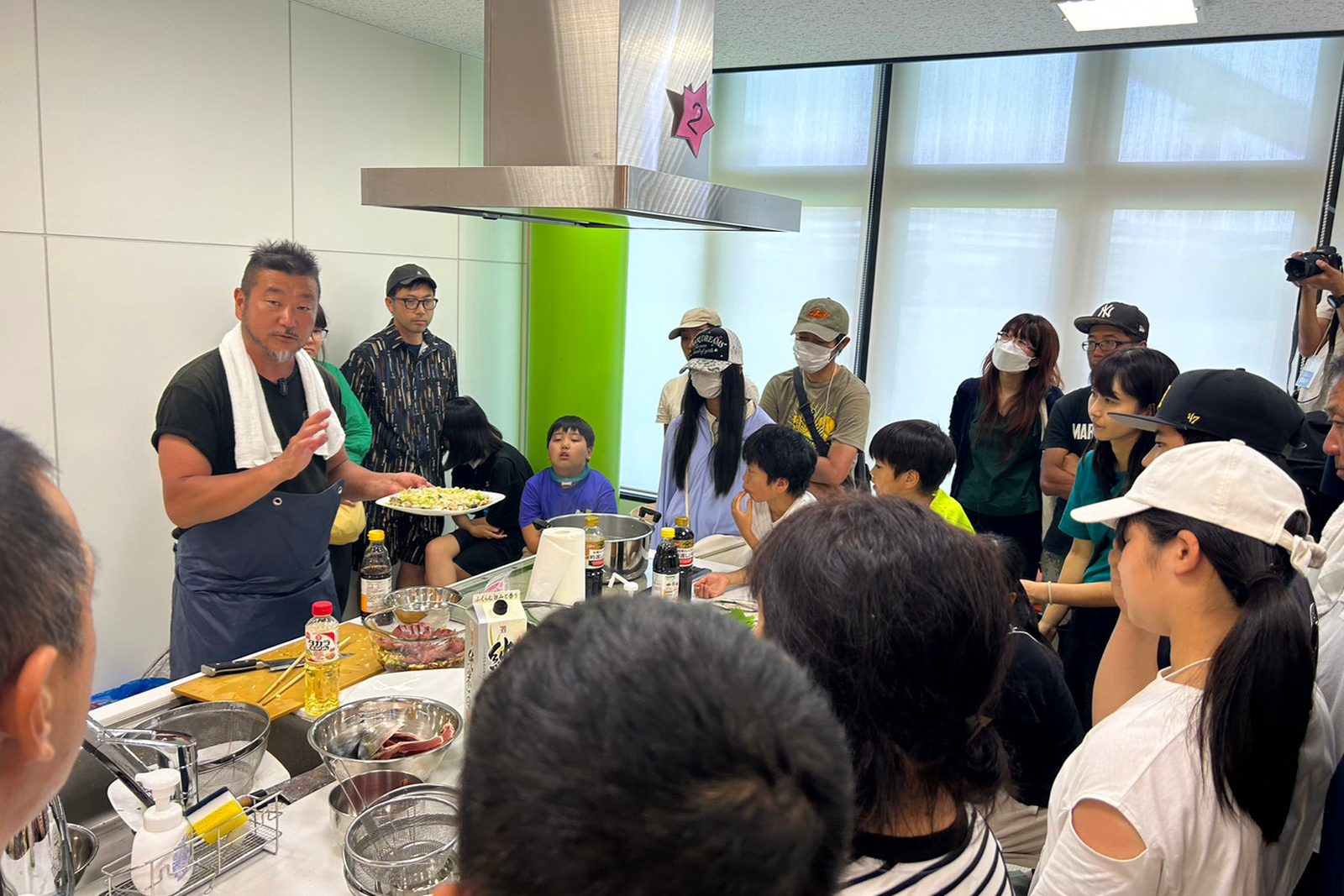 調理台を囲む参加者に、講師が完成した料理を見せている様子