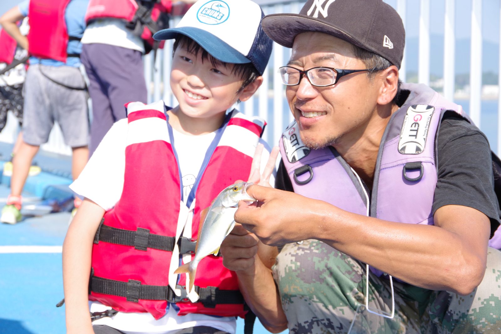 ライフジャケットを着た子どもと、釣れた魚を手にした男性が並んで笑っている